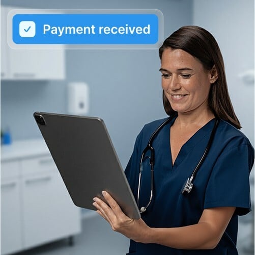A female healthcare professional in blue scrubs is holding a tablet and smiling